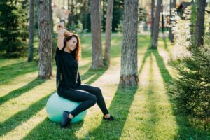 Woman sits on fitness ball raises arms and does excercises outdoor in forest.